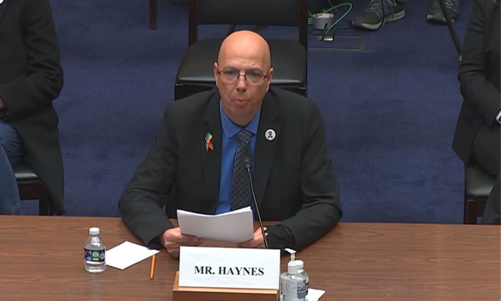 Matthew Haynes prend la parole à une table en bois lors d'un témoignage devant le comité anti-LGBTQ+ sur le discours de haine, assis de chaque côté d'autres témoins et tenant un morceau de papier.