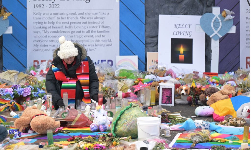 Un individu portant un manteau, une écharpe et un chapeau blanc est assis au milieu des divers hommages rendus à ceux qui sont morts dans la fusillade du Club Q.