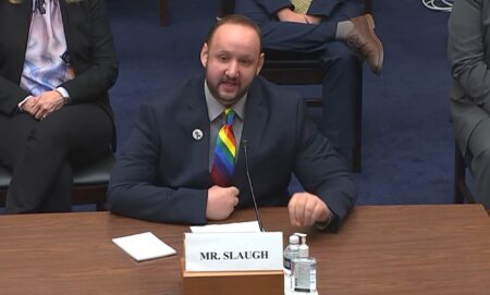 James Slaugh, wearing a rainbow tie and black blazer, speaks to a committe while several people sit behind him during a scheduled meeting.