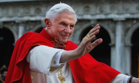 Pope Benedict XVI pictured during his reign wearing white and red robes and raising his hand in a wave to adoring crowds.