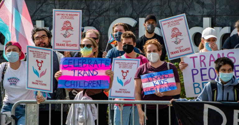 Les membres d'une manifestation pro-LGBTQ+ agitent des signes trans-positifs, ainsi que le drapeau trans rose, bleu et blanc.