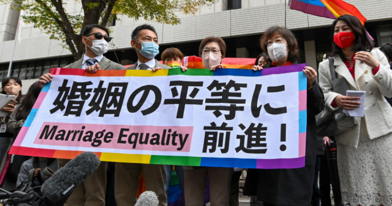 Les plaignants brandissent une bannière sur le thème de l'arc-en-ciel soutenant l'égalité du mariage alors qu'un drapeau arc-en-ciel flotte en arrière-plan.