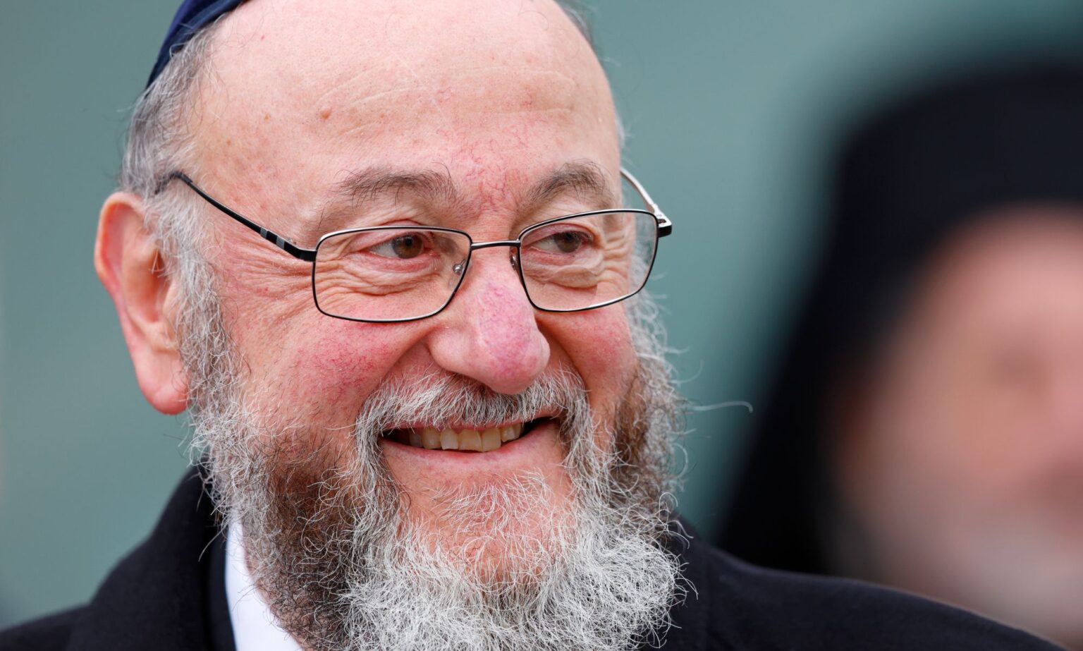 Le grand rabbin du Royaume-Uni défend la communauté LGBTQ+ après le contrecoup du président gay de la Knesset : « Aimez chaque juif » Ephraim Mirvis smiles as he looks off to the left, wearing a black coat.