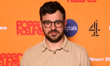 Simon Bird, wearing a cream top, smiles against an orange backdrop.