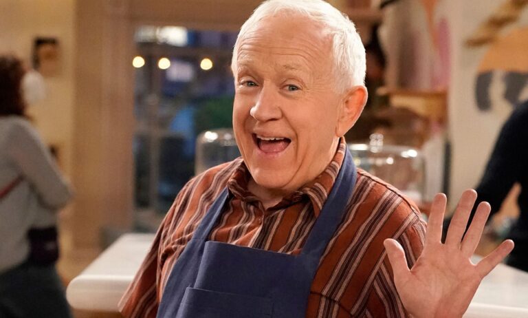 Leslie Jordan smiles and waves while wearing a red striped shirt and baking apron on the set of Call Me Kat