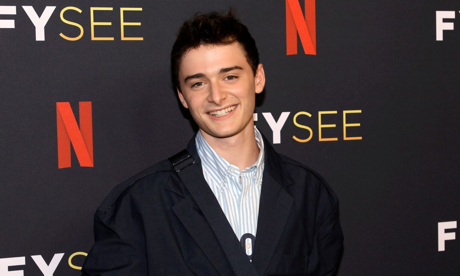 La star de Stranger Things, Noah Schnapp, et ses amis se moquent de son coming-out – et c’est hilarant Noah Schnapp smiles while wearing a light coloured shirt and dark coloured jacket while standing in front of a background with the Netflix logo on it