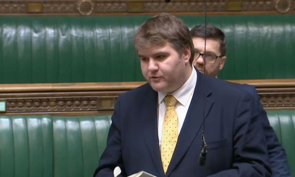 Jamie Wallis porte une chemise blanche, une cravate jaune et une veste bleue lorsqu'il s'adresse au Parlement