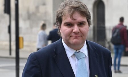 Jamie Wallis wears a white shirt, light blue tie and dark blue jacket while standing outside