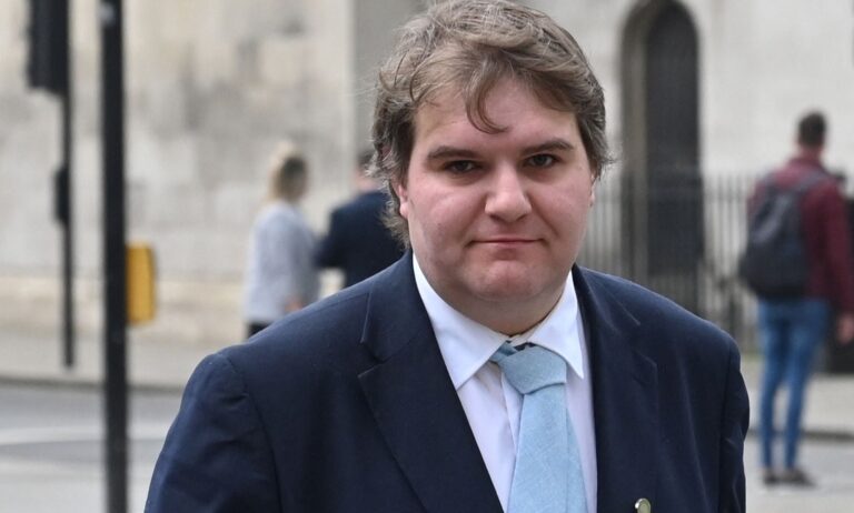 Jamie Wallis wears a white shirt, light blue tie and dark blue jacket while standing outside