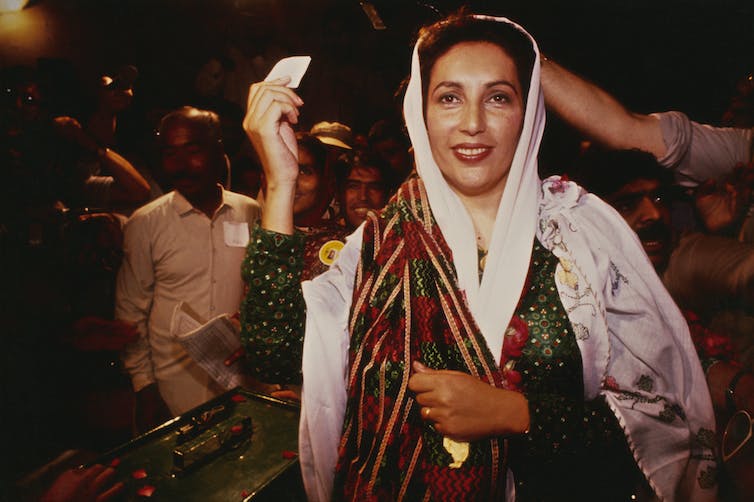 Une femme à la peau brune porte un foulard et des vêtements fluides et tient un petit morceau de papier blanc.