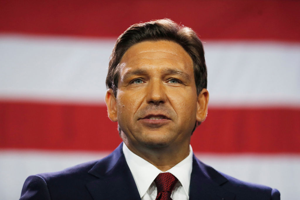 Une photo du gouverneur de Floride Ron DeSantis portant un costume sombre, une chemise blanche et une cravate rouge debout devant le drapeau américain