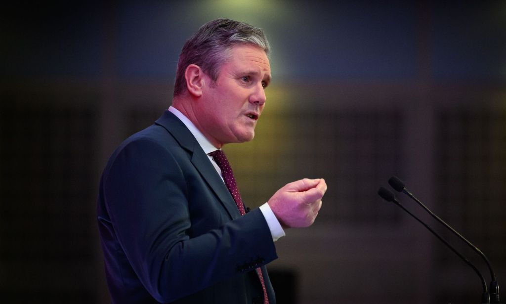 Le chef du Parti travailliste britannique, Keir Starmer, porte une chemise boutonnée blanche, une cravate rouge et une veste bleue lorsqu'il prend la parole lors d'un événement.  Il est debout sur un podium et fait des gestes avec une main tendue