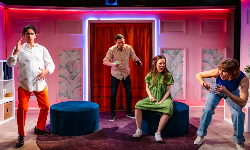 Une photo de la pièce The Boys are Kissing avec les acteurs Seyan Sarvan, Philip Correia, Amy McAllister et Eleanor Wyld se produisant dans un ensemble de style salon rose vif et éclairé au néon.