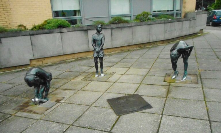 A photo shows the Gorbals Boys artwork piece in Glasgow. There are three statues showing three boys in different positions wearing women's shoes