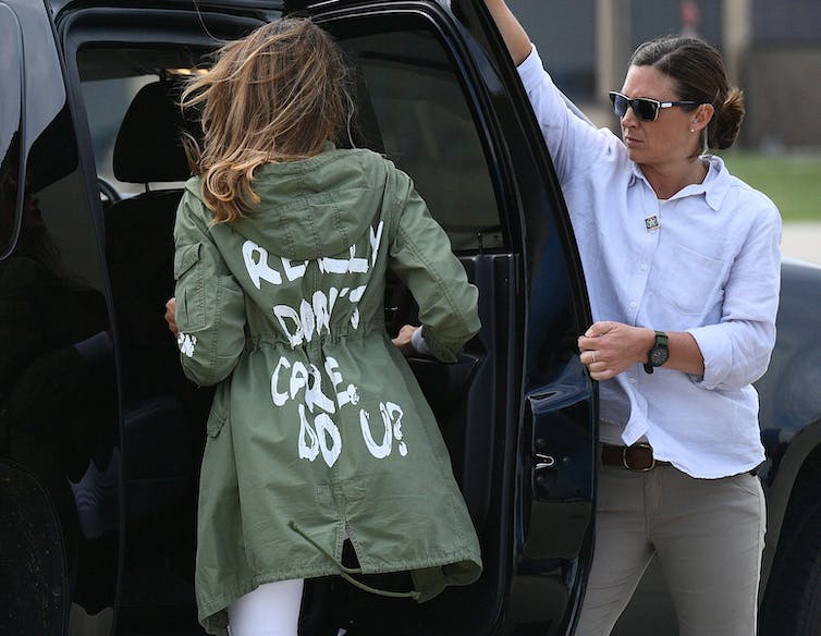 Le dos d'une femme aux longs cheveux bruns monte dans une voiture noire.  Elle porte une veste vert armée avec une écriture blanche.  Une femme vêtue d'une chemise blanche lui tient la porte de la voiture ouverte.
