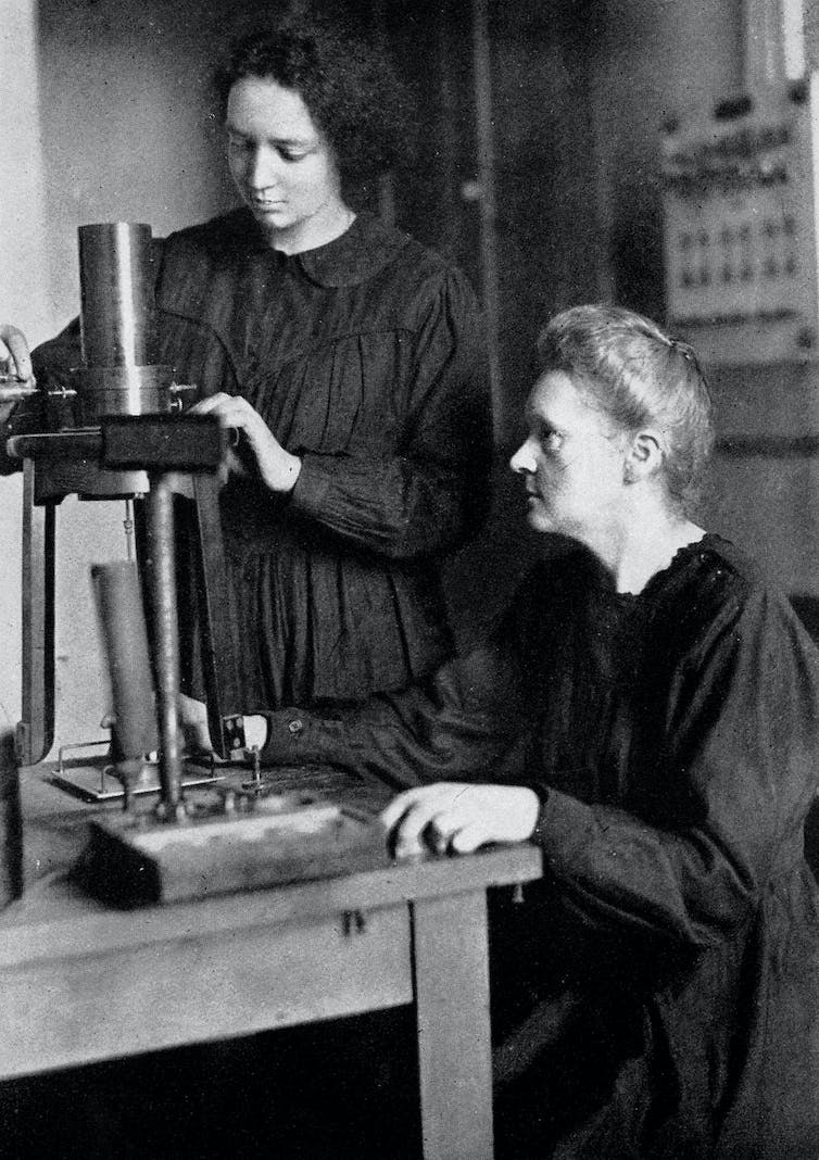 Image en noir et blanc de Marie Curie assise devant une table de travail regardant sa fille régler un instrument.