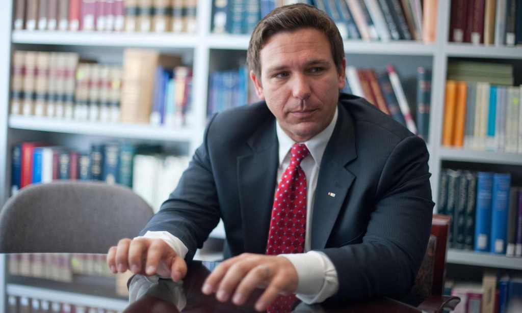 Une photo du gouverneur de Floride Ron DeSantis vêtu d'un costume sombre, d'une chemise blanche et d'une cravate rouge debout dans une pièce avec en arrière-plan des livres sur des étagères