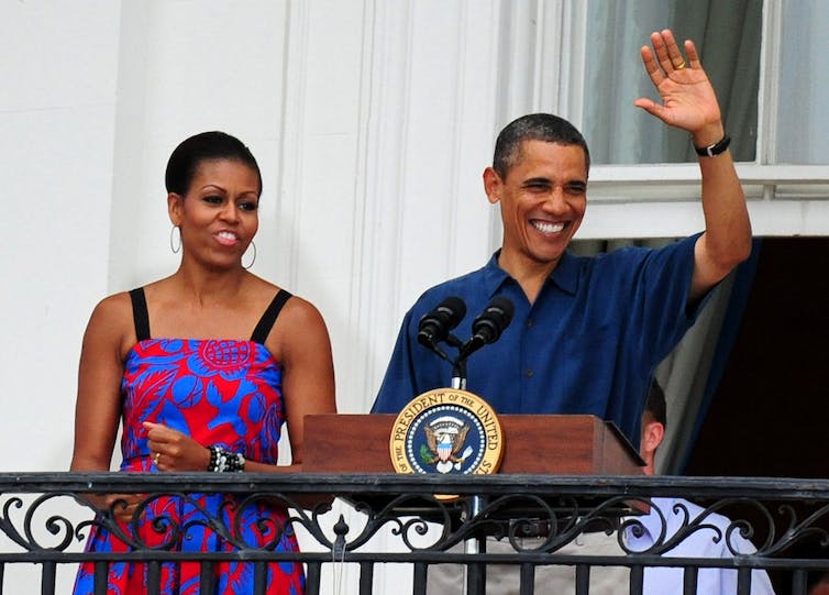 Un homme noir d'âge moyen portant un polo bleu marine salue et sourit sur un balcon, à côté d'une femme noire d'âge moyen avec une robe colorée à motifs rouges et bleus avec de fines bretelles noires.