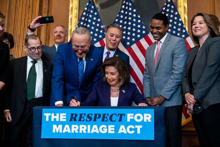 Une femme en blazer signe quelque chose sur une table avec une pancarte indiquant « The Respect for Marriage Act » sous le regard des gens.