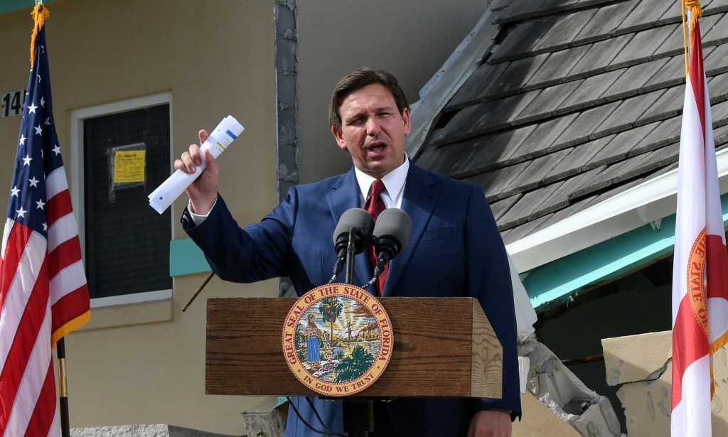 Une photo du gouverneur de Floride Ron DeSantis portant un costume bleu marine, une chemise blanche et une cravate rouge s'exprimant devant un podium