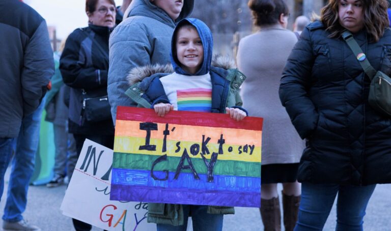 A child in a rainbow T-shirt holds a sign reading "It's okay to say gay"