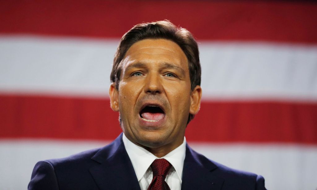 Le gouverneur de Floride, Ron DeSantis, porte un costume et une cravate alors qu'il parle la bouche grande ouverte.  Les rayures rouges et blanches du drapeau américain sont visibles en arrière-plan