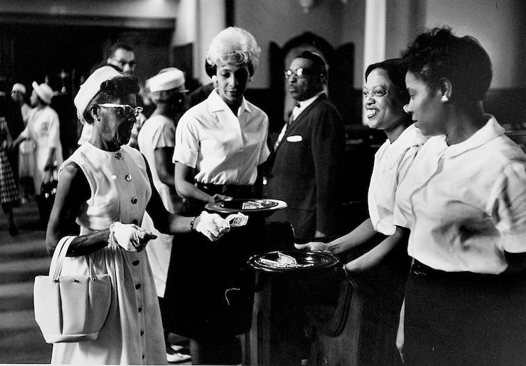 Une photo en noir et blanc montre plusieurs femmes habillées de manière formelle mettant de l'argent dans une assiette de collecte d'église.