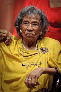 Une femme assise aux cheveux gris vêtue d'une robe et de bijoux dorés.