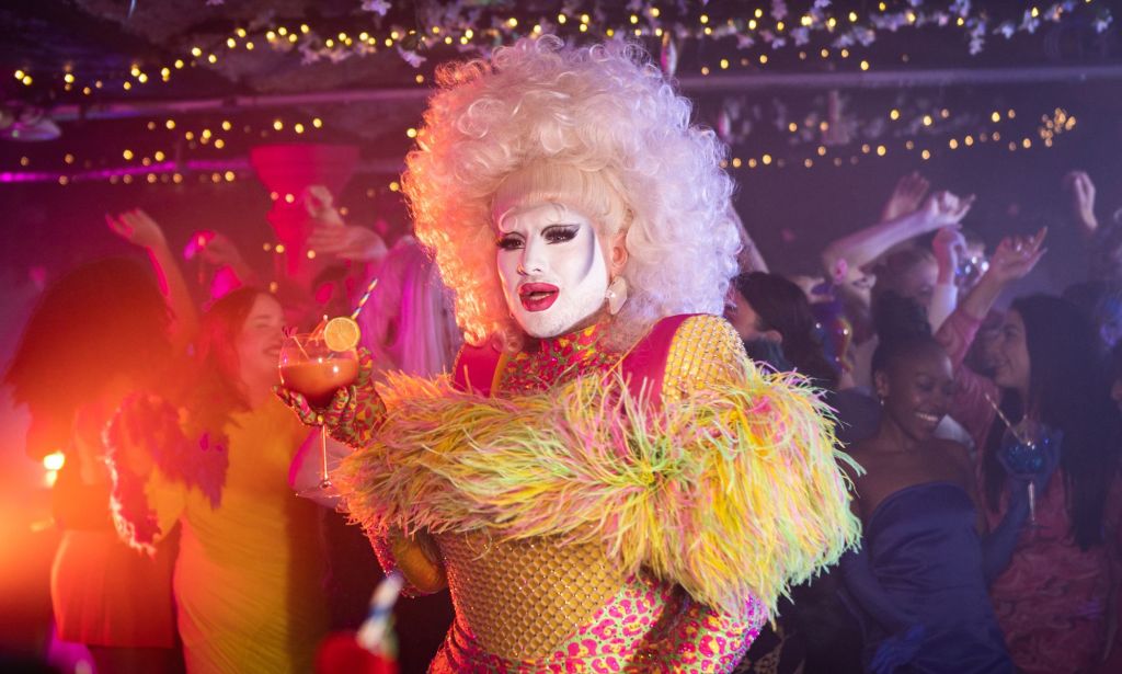 Danny Beard dans un haut jaune et rose moelleux et une perruque blonde, tenant un cocktail.