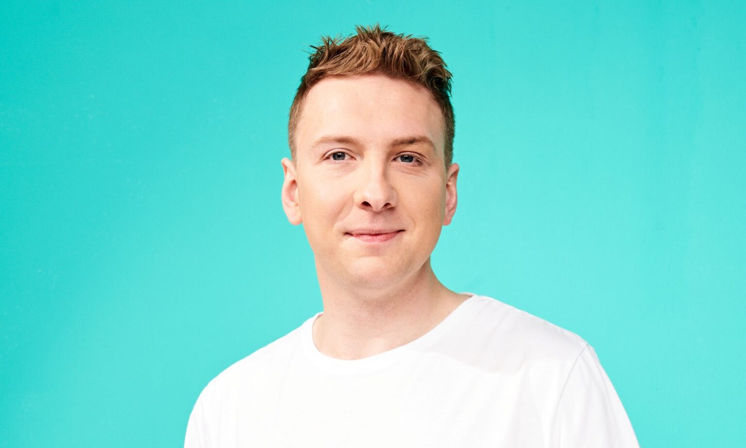 Joe Lycett admet que les cascades virales ont exercé une «pression» sur sa santé mentale – mais il n’est pas prêt à s’arrêter Joe Lycett wearing a white t-shirt and smiling at the camera against a teal background.
