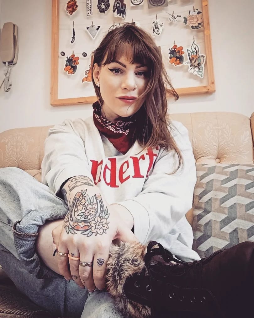 Victoria pose pour la caméra, elle porte un pull blanc avec un bandana et a des tatouages ​​colorés et vifs sur les mains.