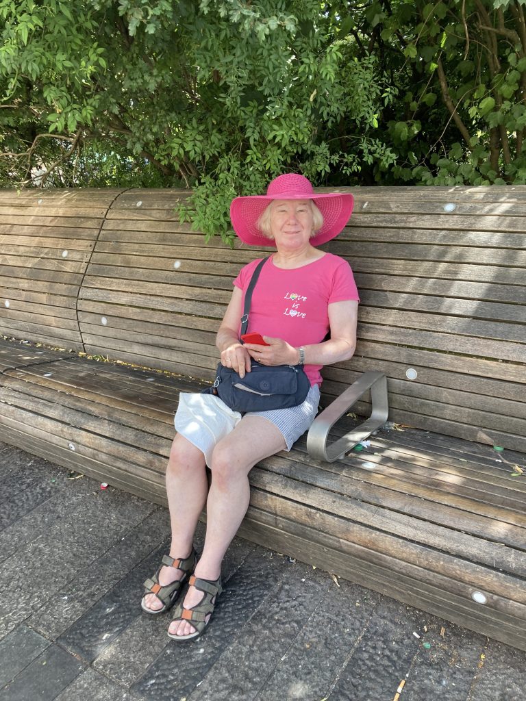 Petra est assise sur un banc en bois.  Elle porte un t-shirt rose et rose et un short en jean. 