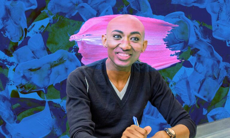 A trans person is smiling for the camera. They are bald, and wearhing a grey v-neck shirt with the sleeves rolled up to their elbows.