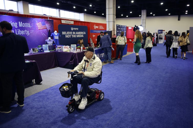 Un homme en fauteuil roulant passe devant un stand dans une salle de congrès qui dit 