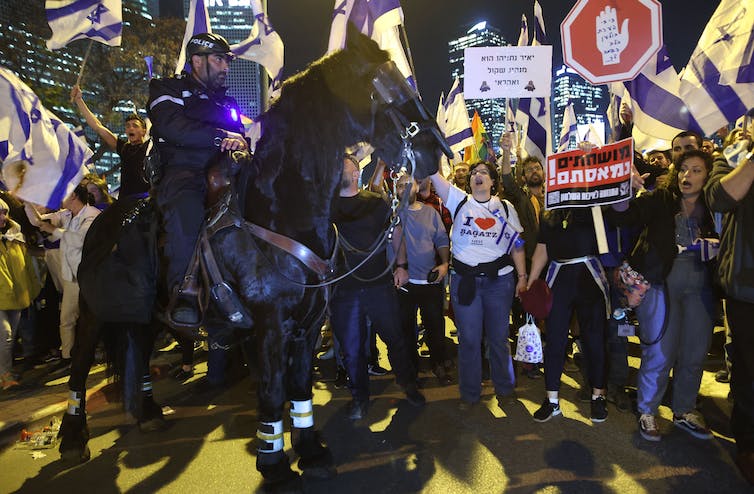 Une foule de personnes portant des pancartes et des drapeaux et criant alors qu'un policier à cheval se tient devant eux pendant la nuit.
