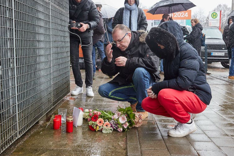 Un homme et une femme en vêtements d'hiver s'accroupissent près d'un mémorial de fortune avec des bougies et des fleurs.