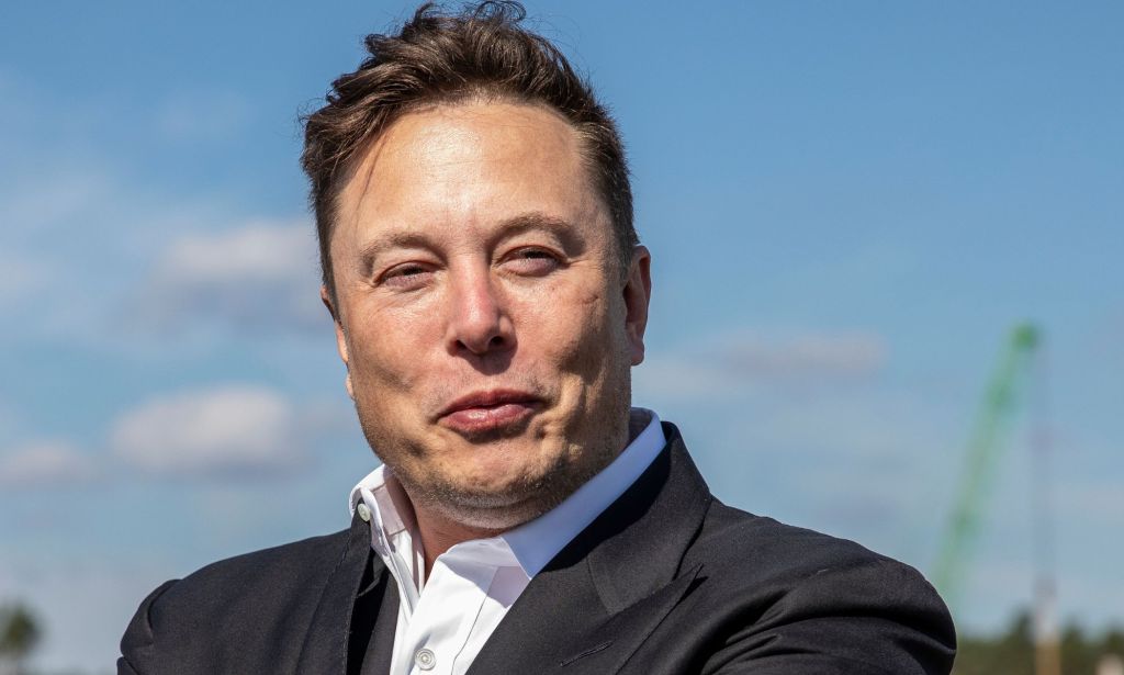 Elon Musk porte une chemise blanche et une veste de costume sombre alors qu'il regarde quelque part hors caméra