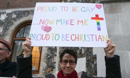 A gay christian holds a sign that reads "proud to be gay, now make me proud to be christian"