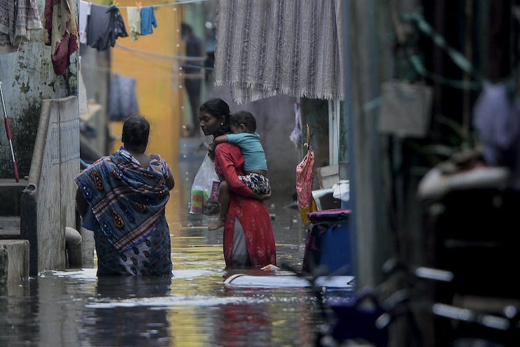 Une femme à la peau brune porte une robe rouge et porte un enfant sur son dos tout en marchant dans l'eau jusqu'aux genoux.
