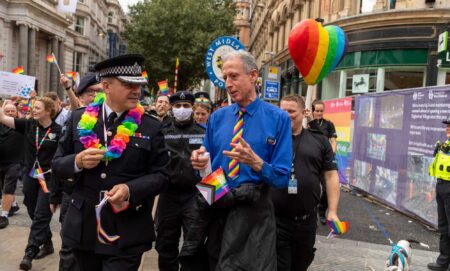 Peter Tatchell talking to the West Midlands Police chief while walking in a Pride march, surrounded by police holding rainbow flags