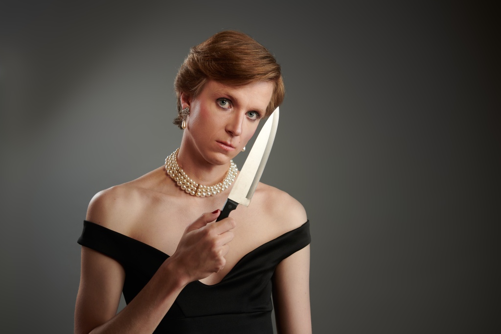Linus Karp dans le rôle de la princesse Diana.  Il porte une robe noire et tient un poignard devant son visage.