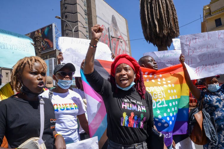 Une manifestation avec une foule de personnes tenant des bannières et des drapeaux arc-en-ciel, devant trois femmes, une avec un poing levé.