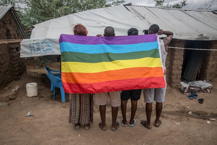 Quatre personnes se tiennent debout, dos à la caméra, tenant un drapeau arc-en-ciel drapé sur leurs épaules.