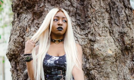 Asexual activist Yasmin Benoit wears a black dress as she poses against a tree and holds part of her blonde hair