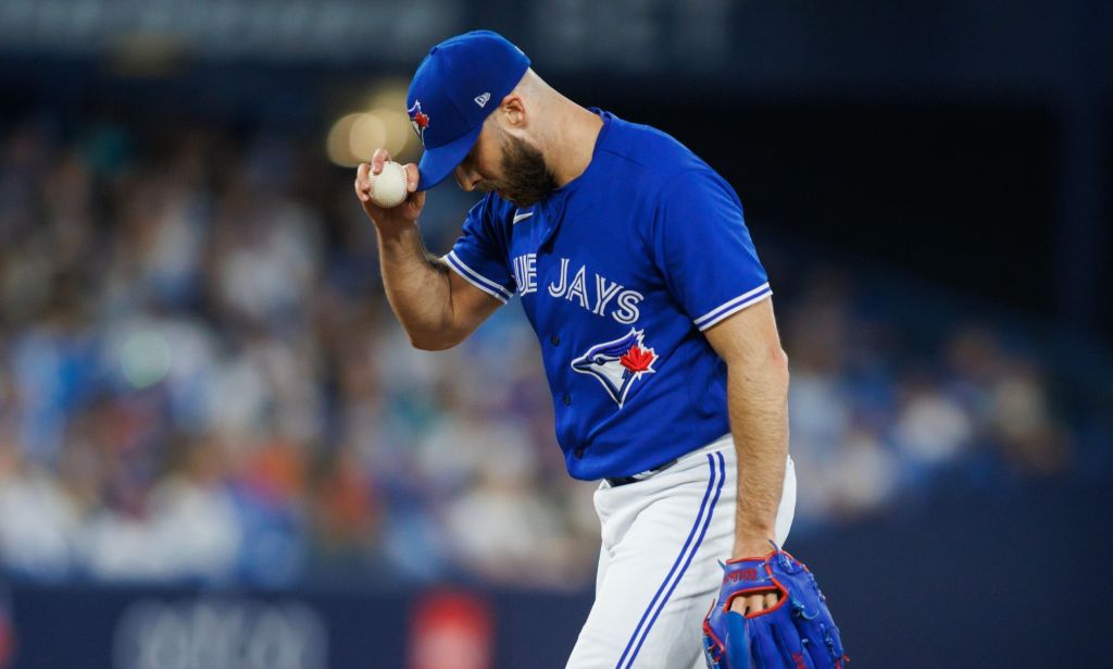 Le lanceur des Blue Jays de Toronto Anthony Bass porte un uniforme de baseball bleu et une casquette assortie avec un pantalon blanc tout en tenant sa casquette et une balle d'une main pendant un match