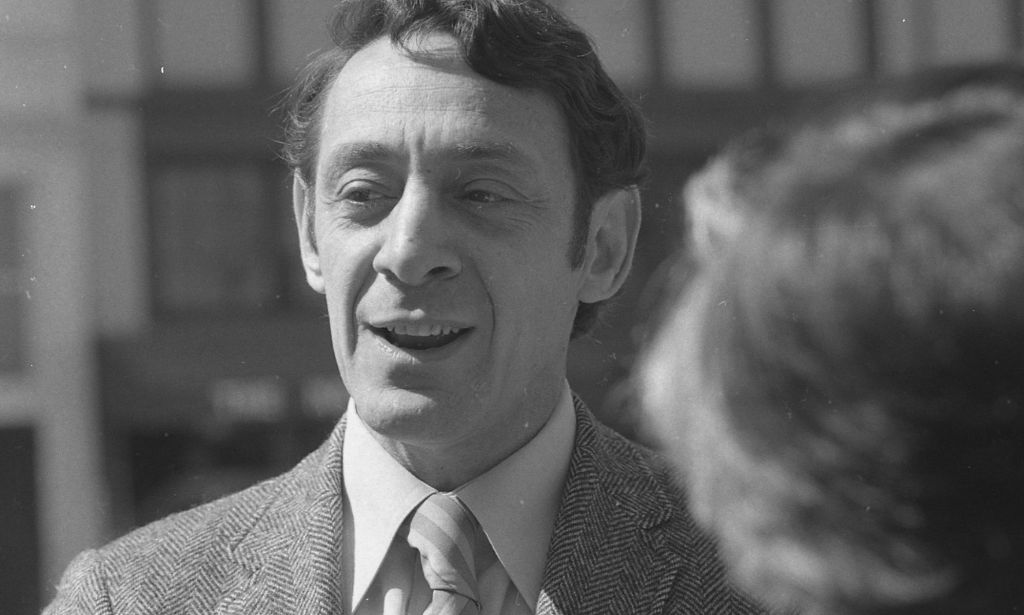 Une photo en noir et blanc de Harvey Milk, qui porte un costume et une cravate, alors qu'il parle aux gens hors caméra