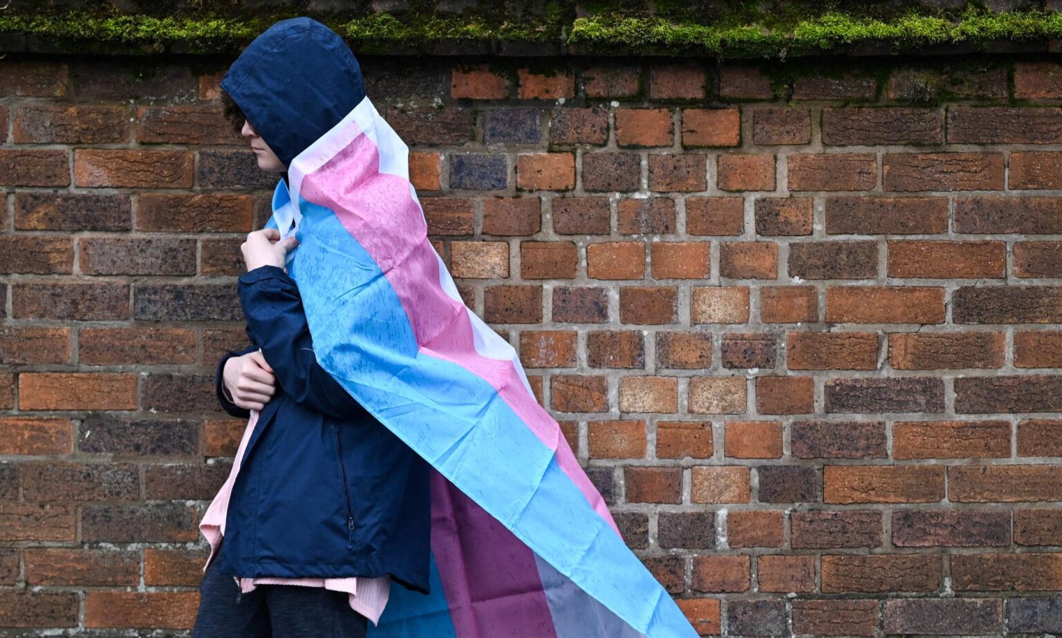 EHRC insiste sur le fait qu’il a «défendu» les droits des trans, malgré les affirmations selon lesquelles il a fait très peu A hooded individual with a trans flag wrapped around them walks in the rain.