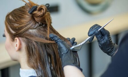 A person with brunette hair sits as someone with a hair dye brush, wearing black gloves, wipes dye on the back of their hair.