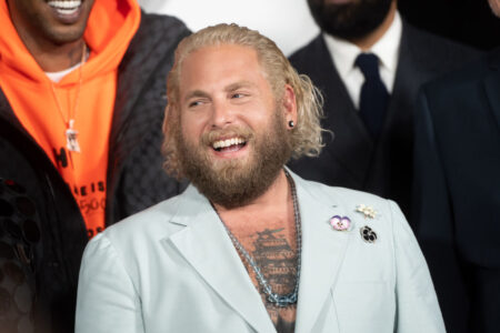 Jonah Hill, a white man with long bleached hair, wearing a light blue suit reveaing his tattooed chest