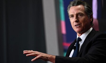 California governor Gavin Newsom wears a suit and tie as he gestures off screen, he recently threatened to fine a school district for not using materials mentioning Harvey Milk